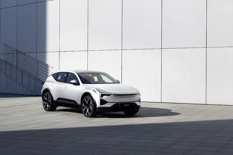 White Polestar 3 electric SUV parked on plaza in front of minimalist white panel wall in bright sunlight, front three-quarter view