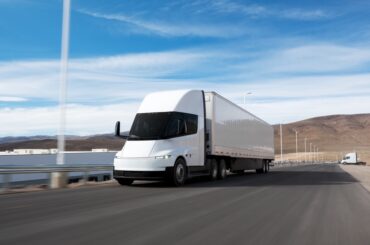 White semi-truck driving on a straight desert highway under a clear blue sky with distant hills.