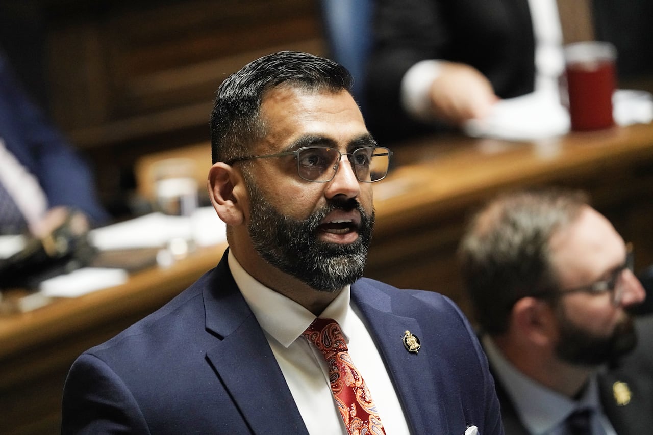 A man in a blue suit and reddish tie speaks during a government legislative session.