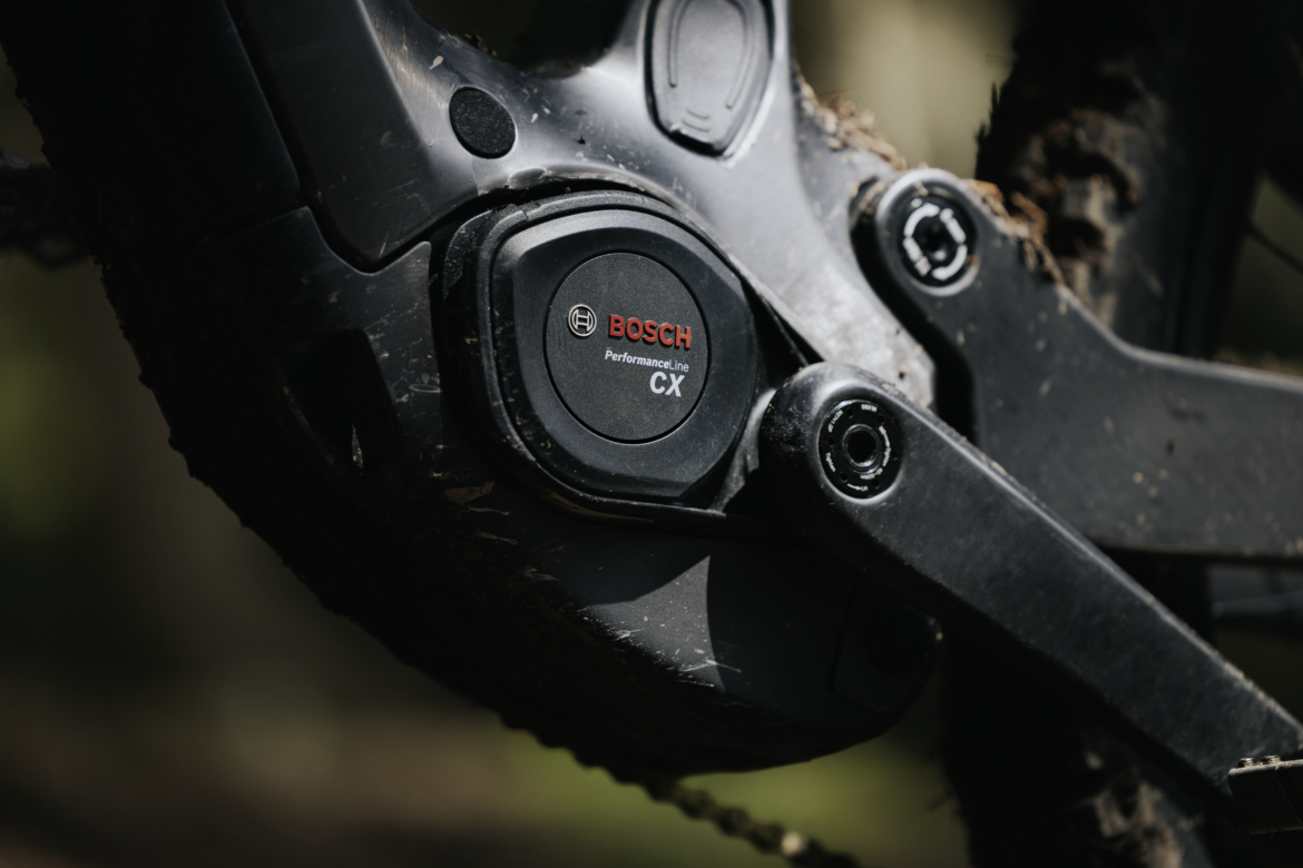 Close-up view of a Bosch Performance Line CX motor mounted on a mountain bike, showing the casing and surrounding components, with dirt and mud visible, indicating outdoor use.