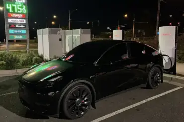 Black Tesla Model Y plugged into a Tesla Supercharger at night with nearby gas prices displayed in the background.