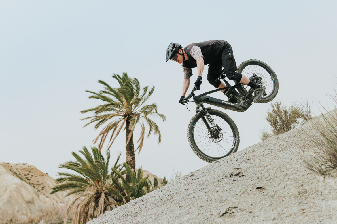 A person riding a mountain bike performs a trick on a sandy slope, with a backdrop of palm trees and a clear sky. The rider is wearing a helmet and athletic clothing, focused on maintaining balance while navigating the terrain.