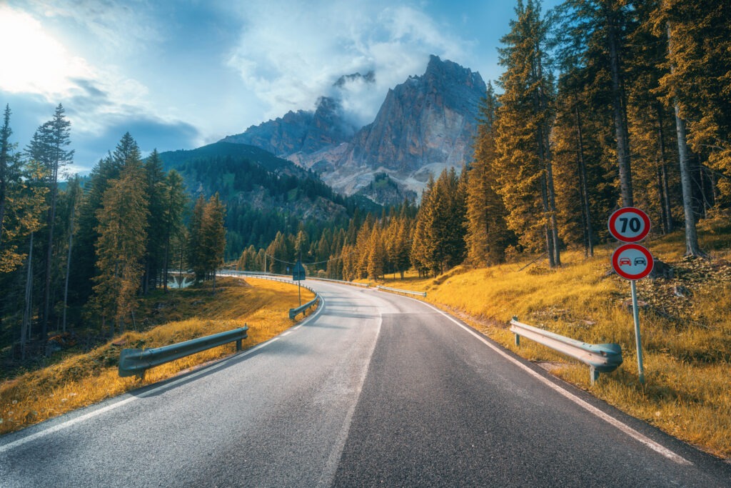 Road in alpine mountains at sunset in autumn. Dolomites, Italy. Beautiful road, orange tress, high rocks in clouds. Landscape with empty highway through the mountain pass in fall.