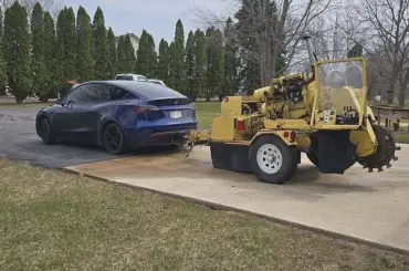 Blue Tesla Model Y towing a yellow construction trencher on a small trailer in a parking lot.