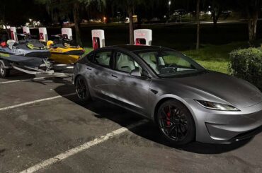 Tesla Model 3 towing two Sea-Doo personal watercraft on a trailer at a Tesla Supercharger station at night.
