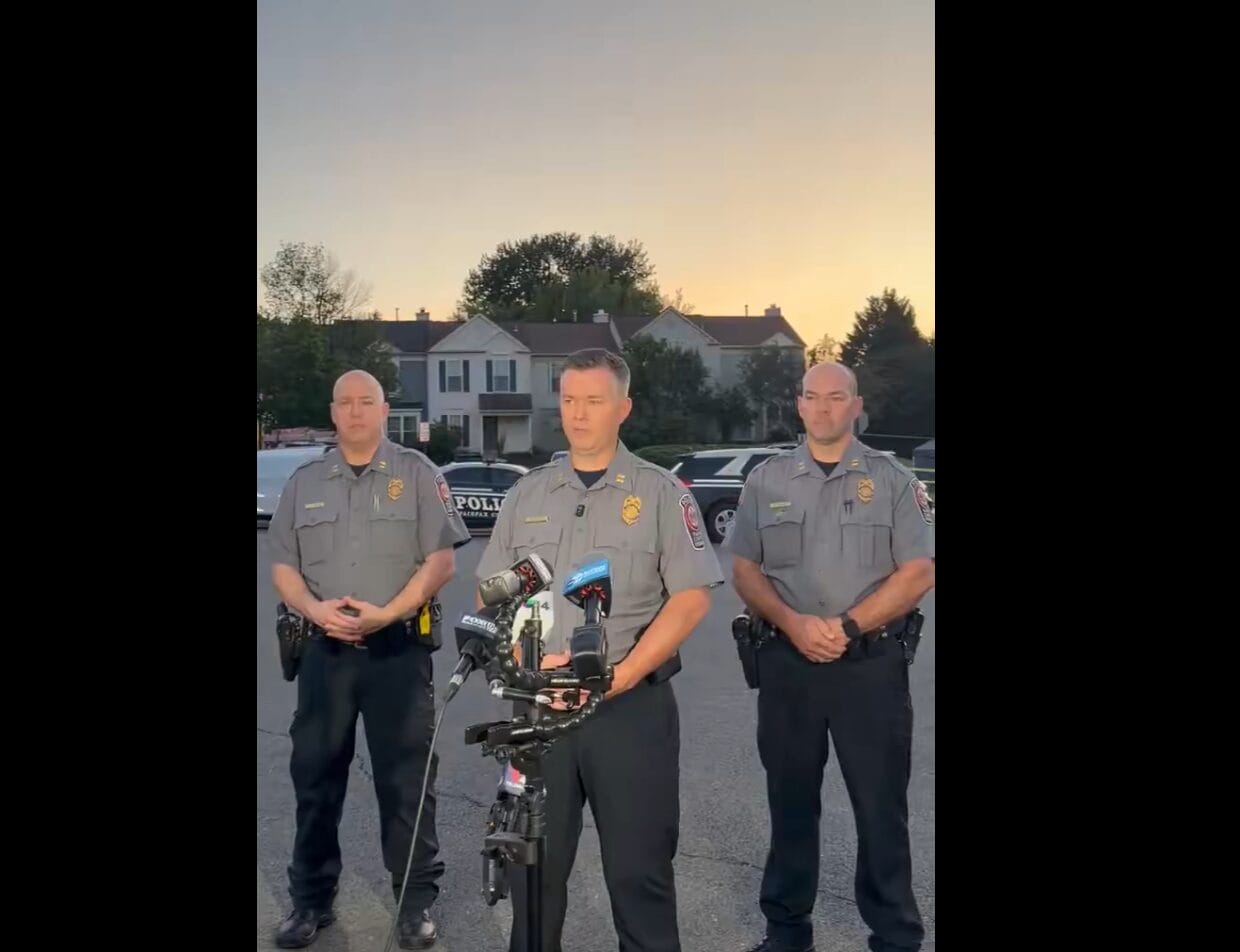 Three uniformed police officers stand in a parking lot at sunset, microphones in front of the center officer as he speaks.