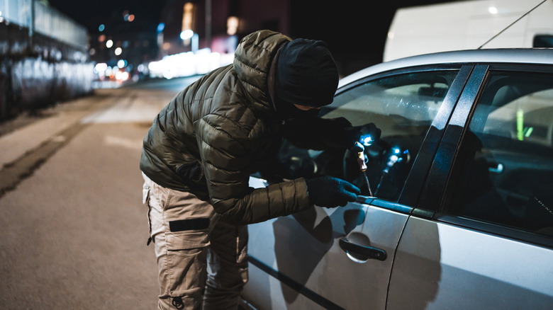 A person wearing a green hoodie and a black ski mask trying to break into a car at night