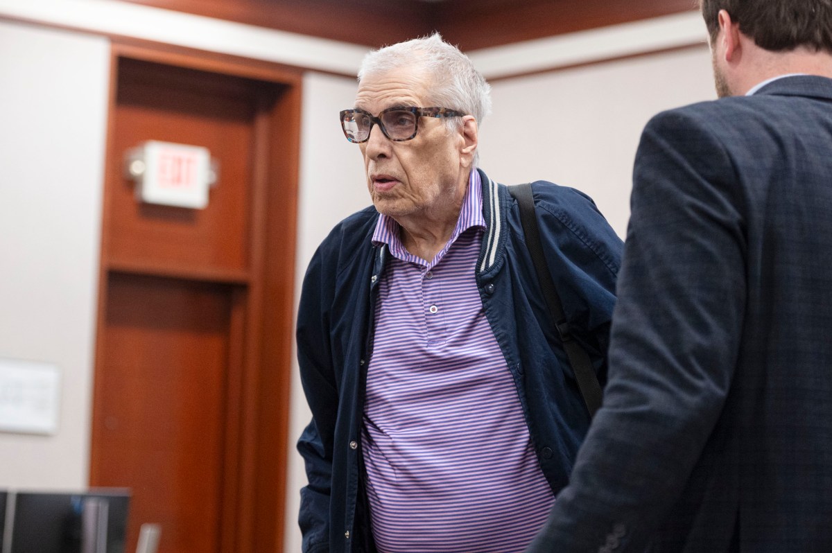 Older man wearing glasses and a striped polo shirt stands beside another man in a suit in a courtroom setting.
