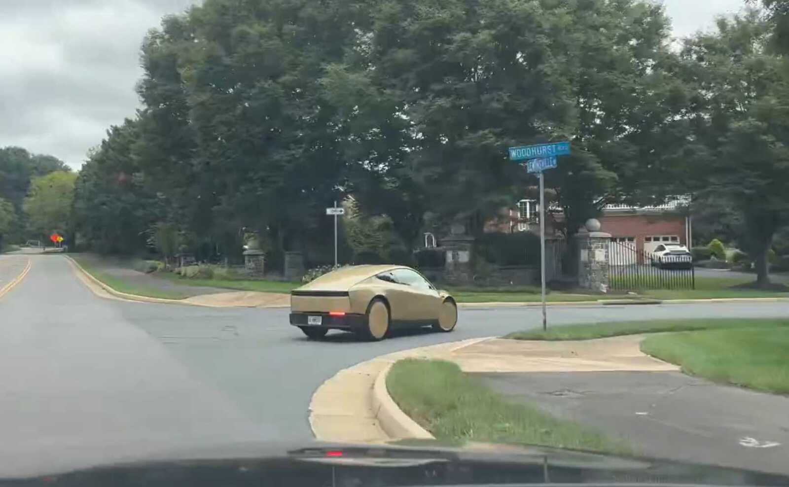 Report: Self-driving taxi from Tesla spotted on Fairfax County roads Beige, bubble-shaped concept car turning at a suburban intersection with trees and houses in the background.
