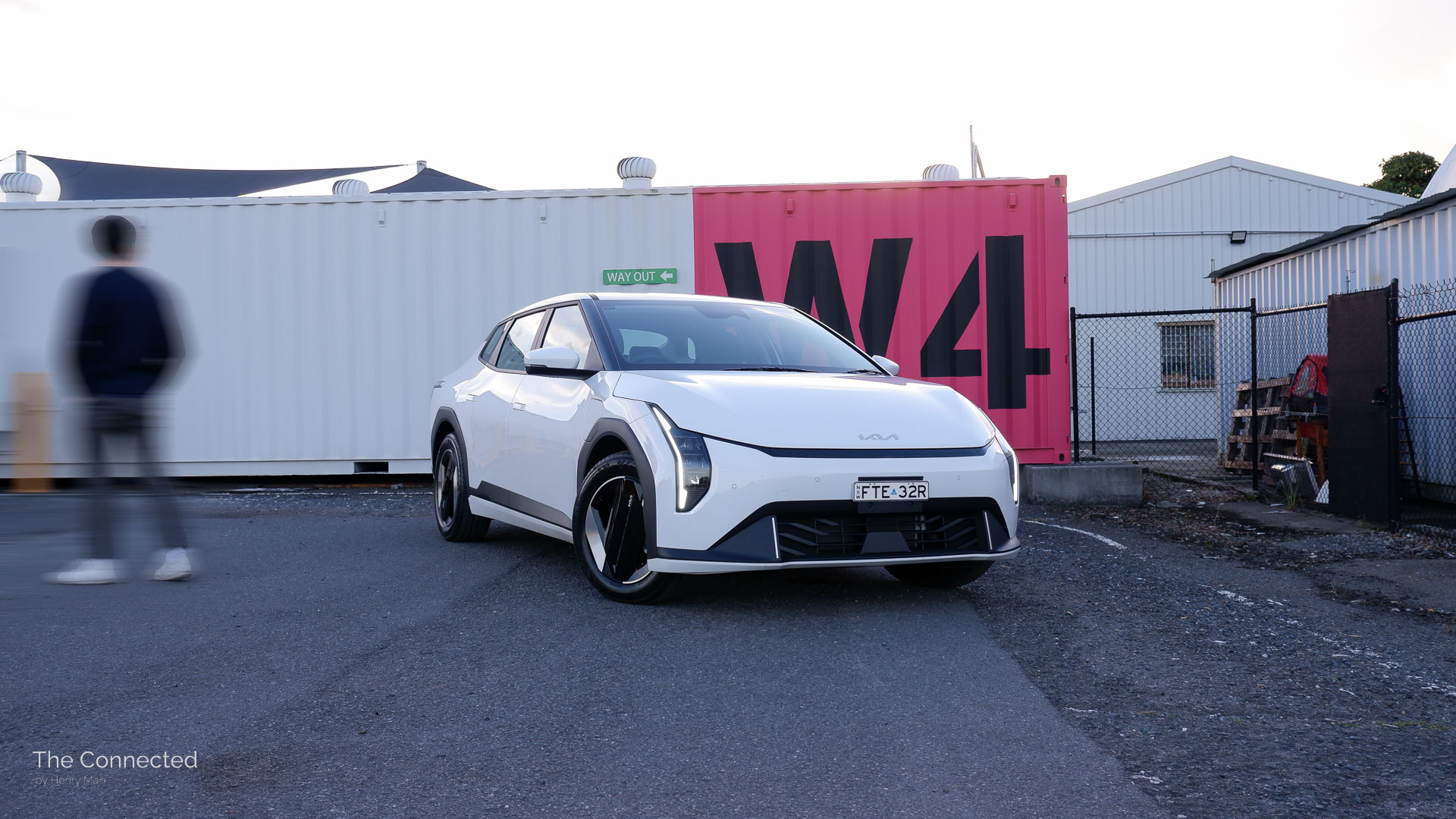 Man walks past the 2026 Kia EV4 Air with a white shipping container behind painted with 'W4' in pink and black