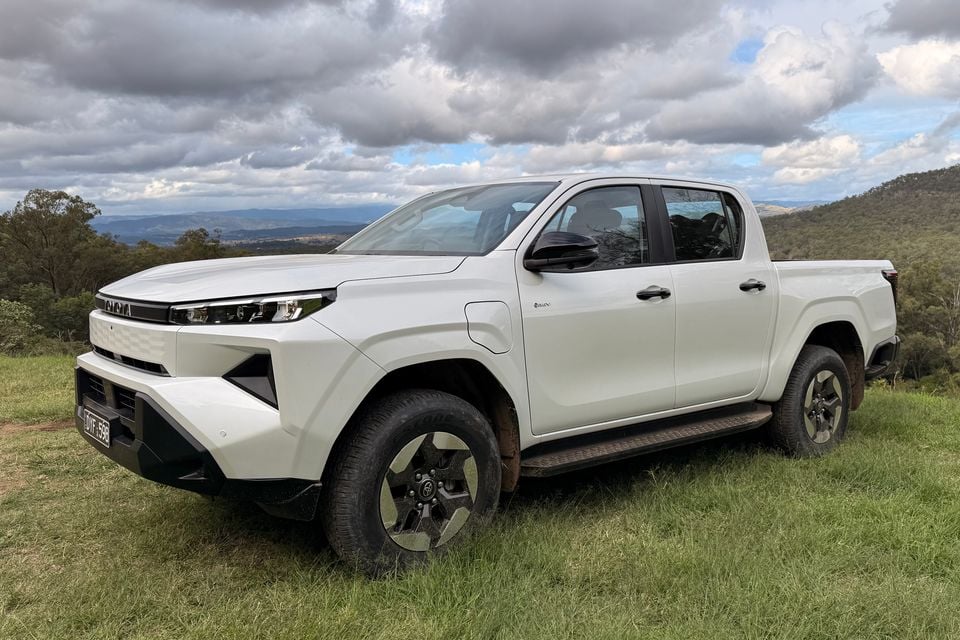 A white Toyota HiLux dual-cab ute parked on a grassy hillside with a scenic mountain backdrop, seen from the front-side angle